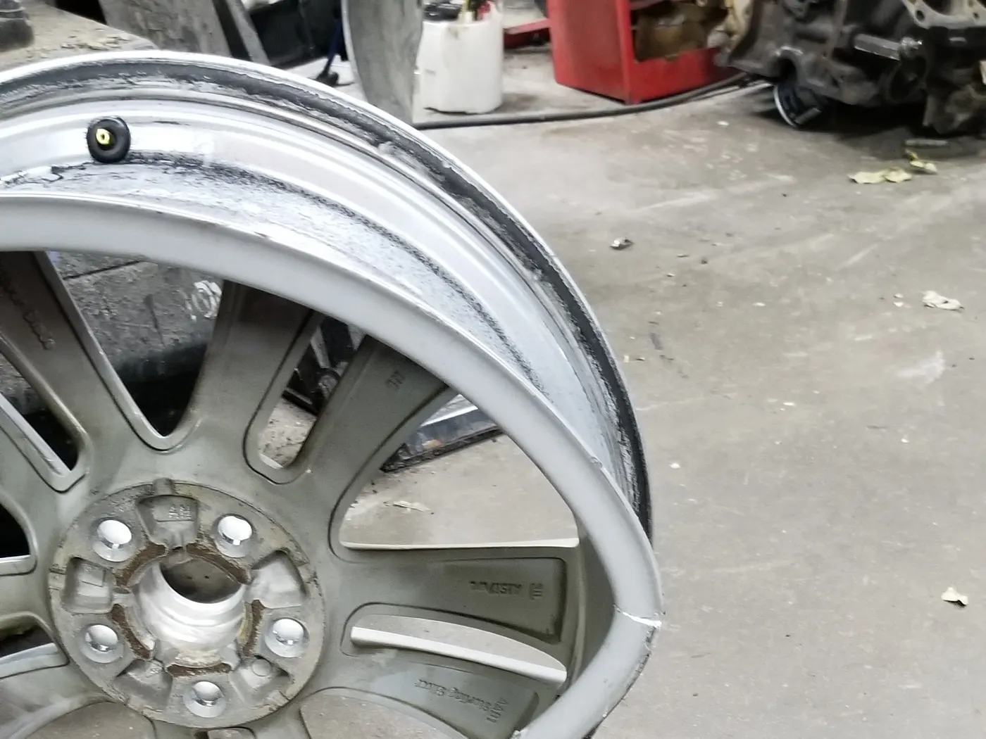 A bent alloy wheel on the shop floor at Ultimate Automotive, an engine block and a red tool cart behind it