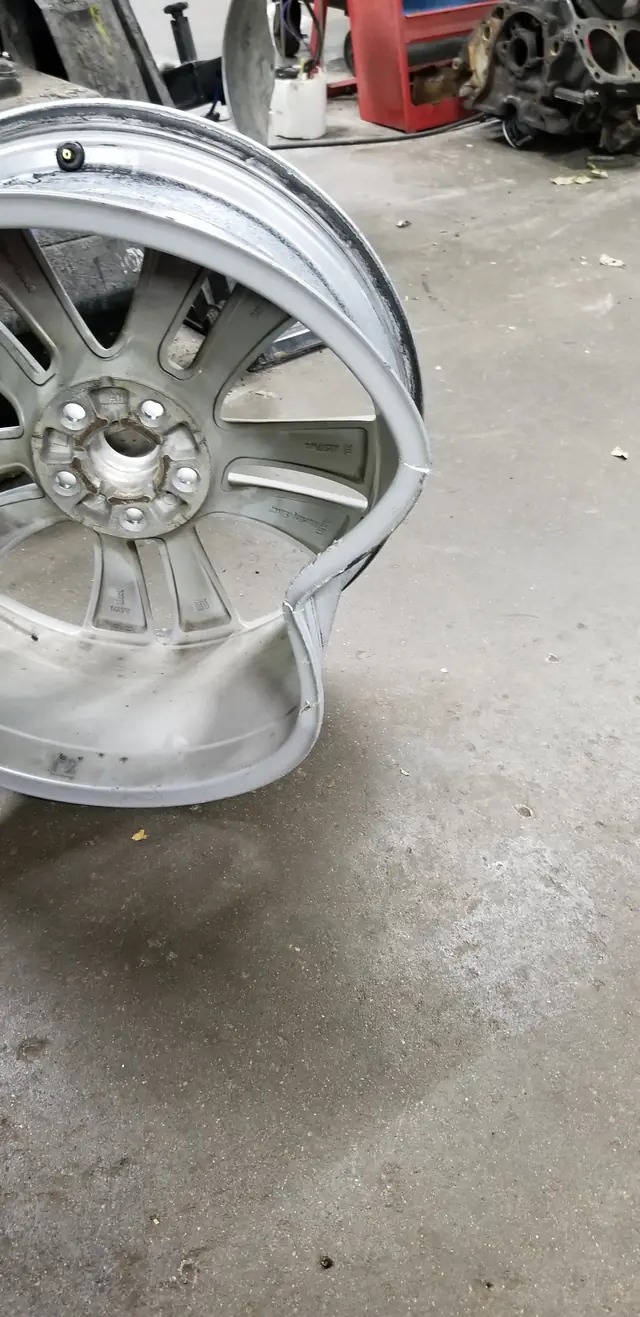 A bent alloy wheel resting on the shop floor with an engine block and a red tool cart behind it