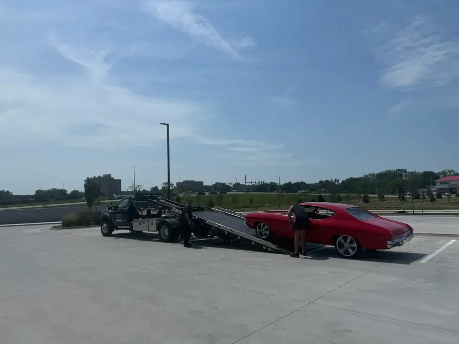 An Ultimate Towing flatbed unloading a red Chevelle in a parking lot under a wide Iowa sky